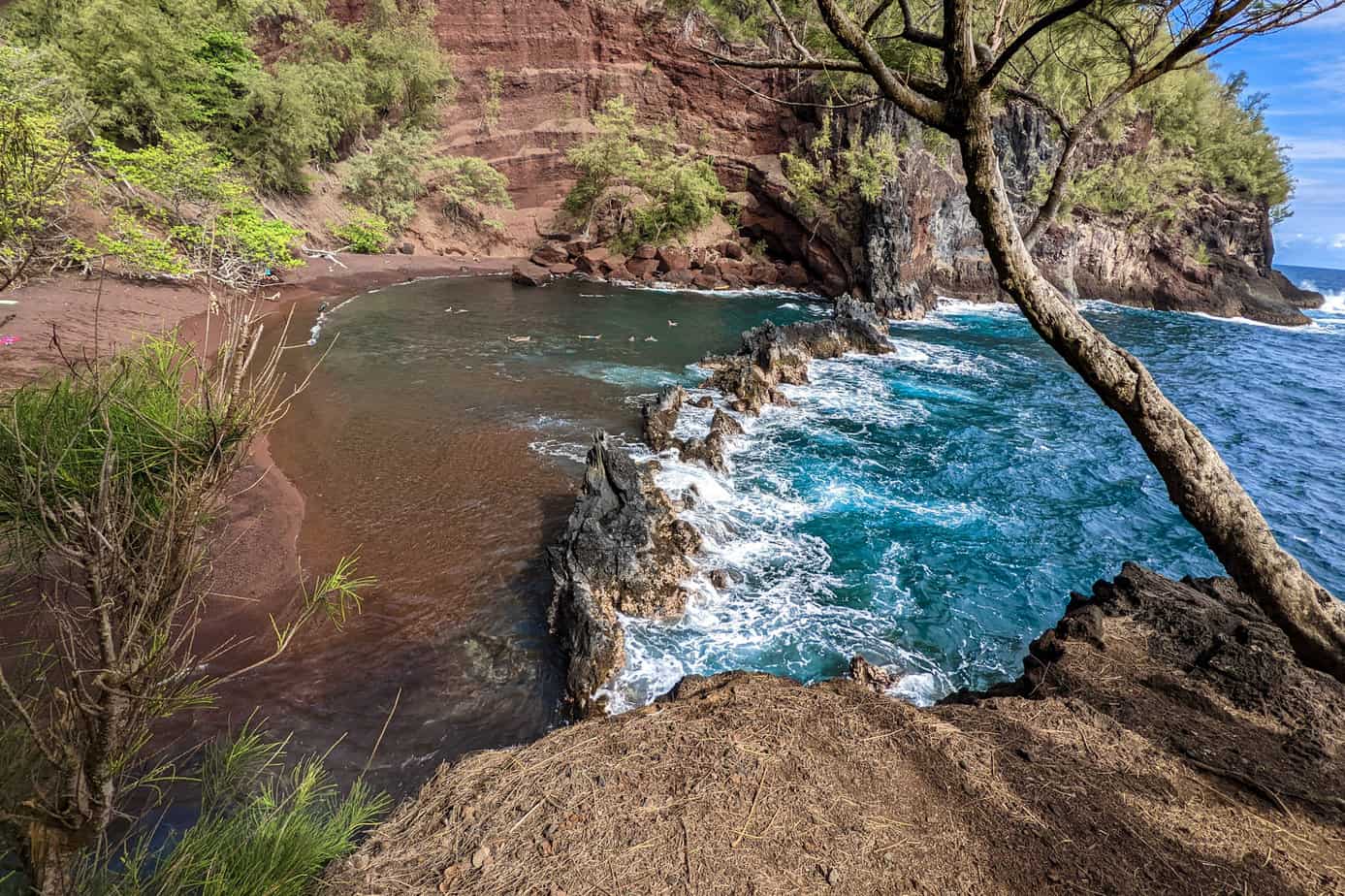 Red sand beach in a cove, protected by black lava rock, blue waves crashing on the protective barrier
