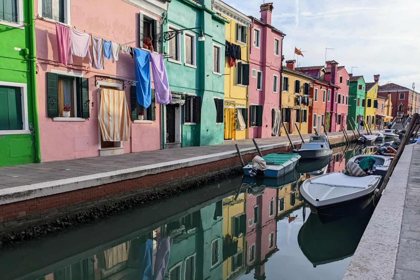 Colorful houses line water in Burano. Boats sit in the canal. Pink, Green, and yellow houses alternate on the water.