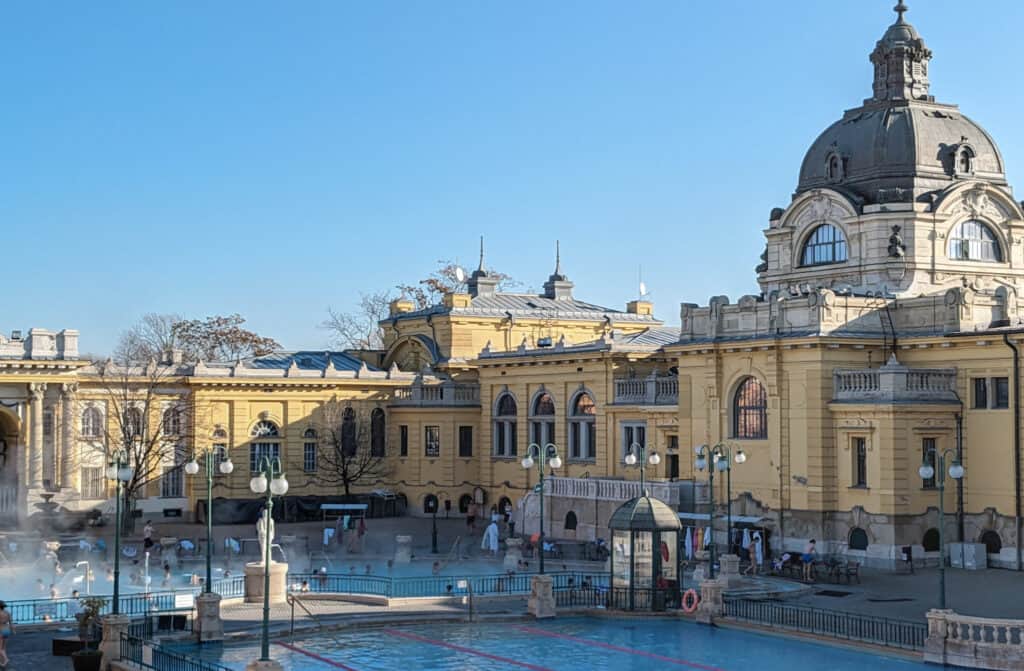 View of the interior of Szechenyi Baths. The yellow strone building with steam from two different exterior thermal baths.