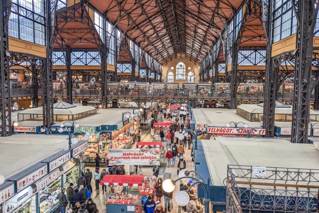 A view of the interior of the Central Market Hall in Budapest, Hungary. The image shows a bustling marketplace filled with vendors selling various fruits, vegetables, meats, and other goods, with shoppers walking among the stalls
