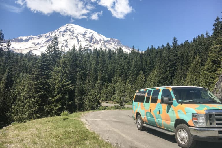 A view of a campervan parked near Mount Rainier in Washington. The image shows the snow-capped peak of the mountain in the background, with a colorful campervan in the foreground.