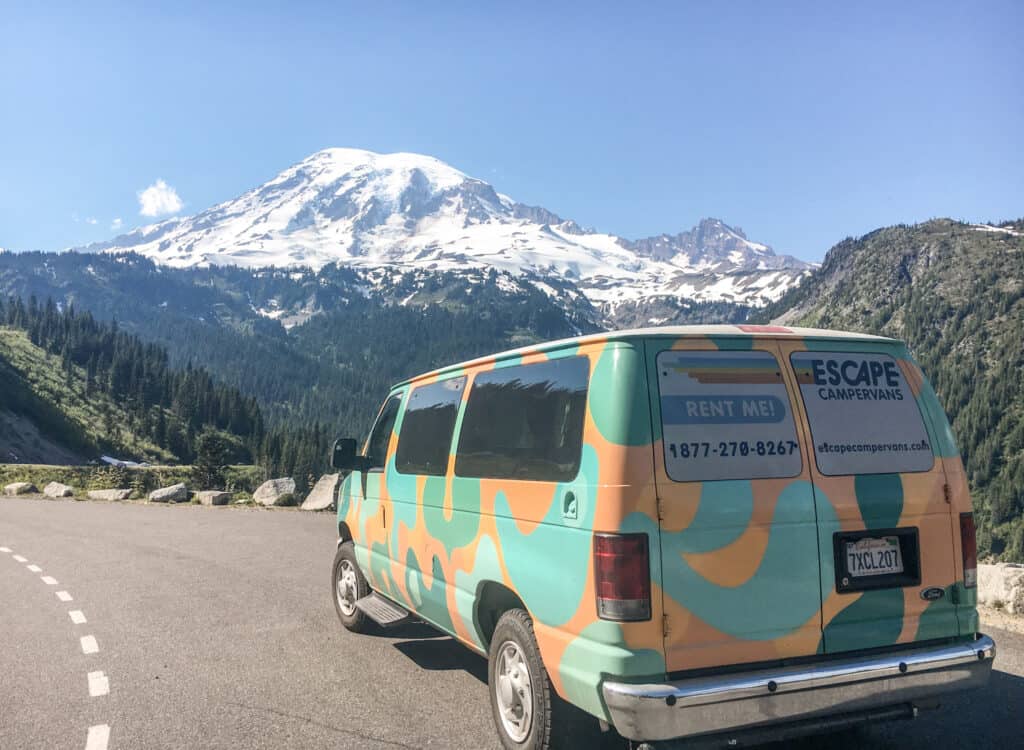 A view of a campervan parked near Mount Rainier in Washington. The image shows the snow-capped peak of the mountain in the background, with a colorful campervan in the foreground.