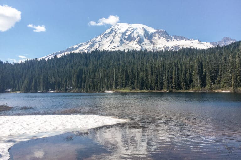 A view of Reflection Lake in Mount Rainier National Park, Washington, with Mount Rainier in the background. The image shows the tranquil lake reflecting the snow-capped peak of the mountain, with trees and foliage visible in the foreground