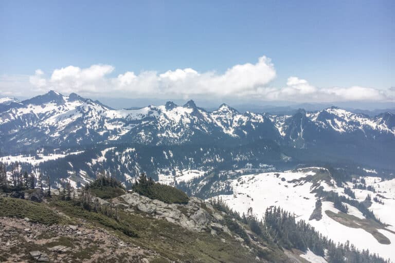 A panoramic view from Panorama Point on Mount Rainier in Washington. The image shows a stunning view of the surrounding mountains and valleys, with clouds and blue sky visible in the distance