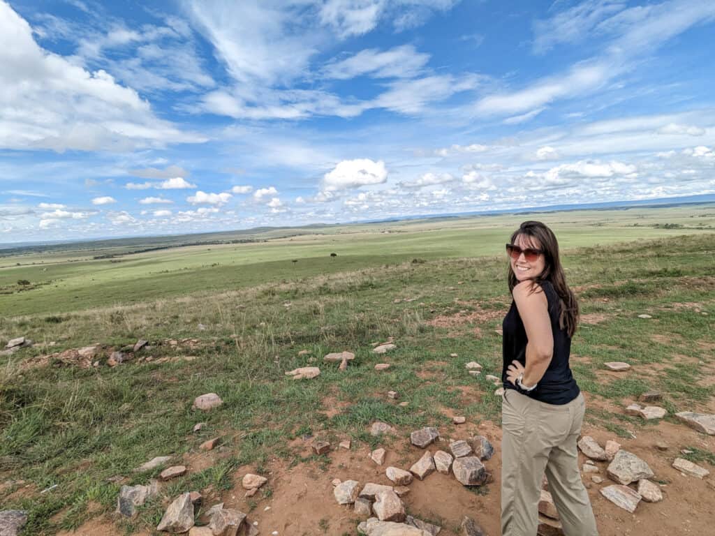 The view from a high point in Masai Mara- Jami stands in the photo. Blue skies and green grasslands in the background.